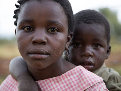 A young girl looks intently at the camera while holding a crying young boy on her back.