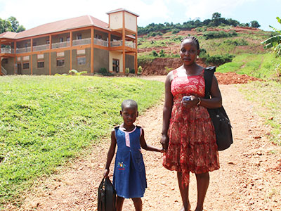 Young girl in a blue dress stands next to her mom, holding her hand, at the Kireka Kinship Project in Uganda