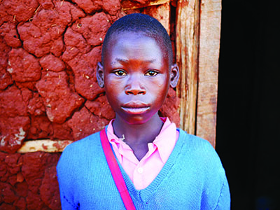 Young girl from Kenya stands in front of a mud wall