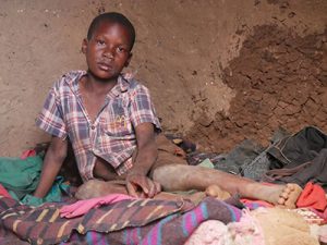 Young boy with dirty clothes sits solemnly on blankets in a mud house.