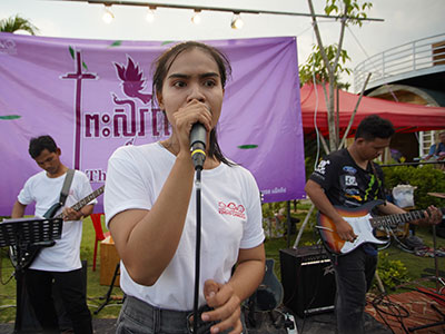 A woman sings into a microphone as two men play guitars behind her