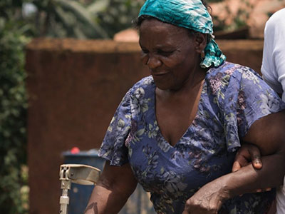 A woman walks into a medical outreach event in Uganda with the help of her crutch and another person
